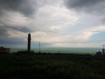 Vor einem Minarett liegt eine Grasfläche im Schatten einer dunklen Regenwolke. Dahinter fällt ein tiefer Berghang ab, wodurch man weit über eine grüne Ebene blicken kann.