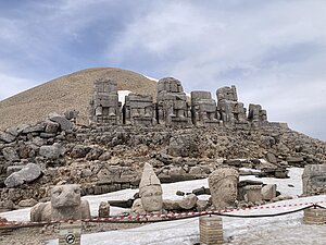 Auf einer Bergspitze stehen sechs Statuen sitzender, anthropomorpher Gestalten; allen fehlt der Kopf, einigen auch größere Teile des Oberkörpers. Die Köpfe (ein Adler und mehrere Gottheiten) stehen in einigem Abstand zu Füßen der Statuen. 