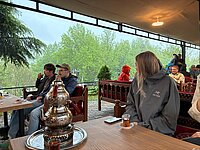 Vor einer verregneten Landschaft sitzt eine Gruppe auf mehreren geschnitzten Holzbänken beim Tee.