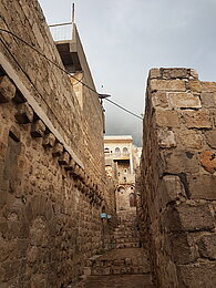 Eine schmale Treppe führt zwischen Steinmauern zu einer hölzernen Haustür. Dahinter erhebt sich ein verzierter Altbau, über dem eine dunkle Regenwolke aufzieht.