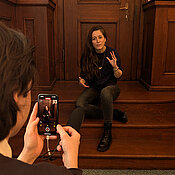 Anne Sauer sitzt auf Holztreppe und erzählt und gestikuliert dabei. Sind wird von einer anderen Person mit einem Smartphone gefilmt und spricht in die Kamera.